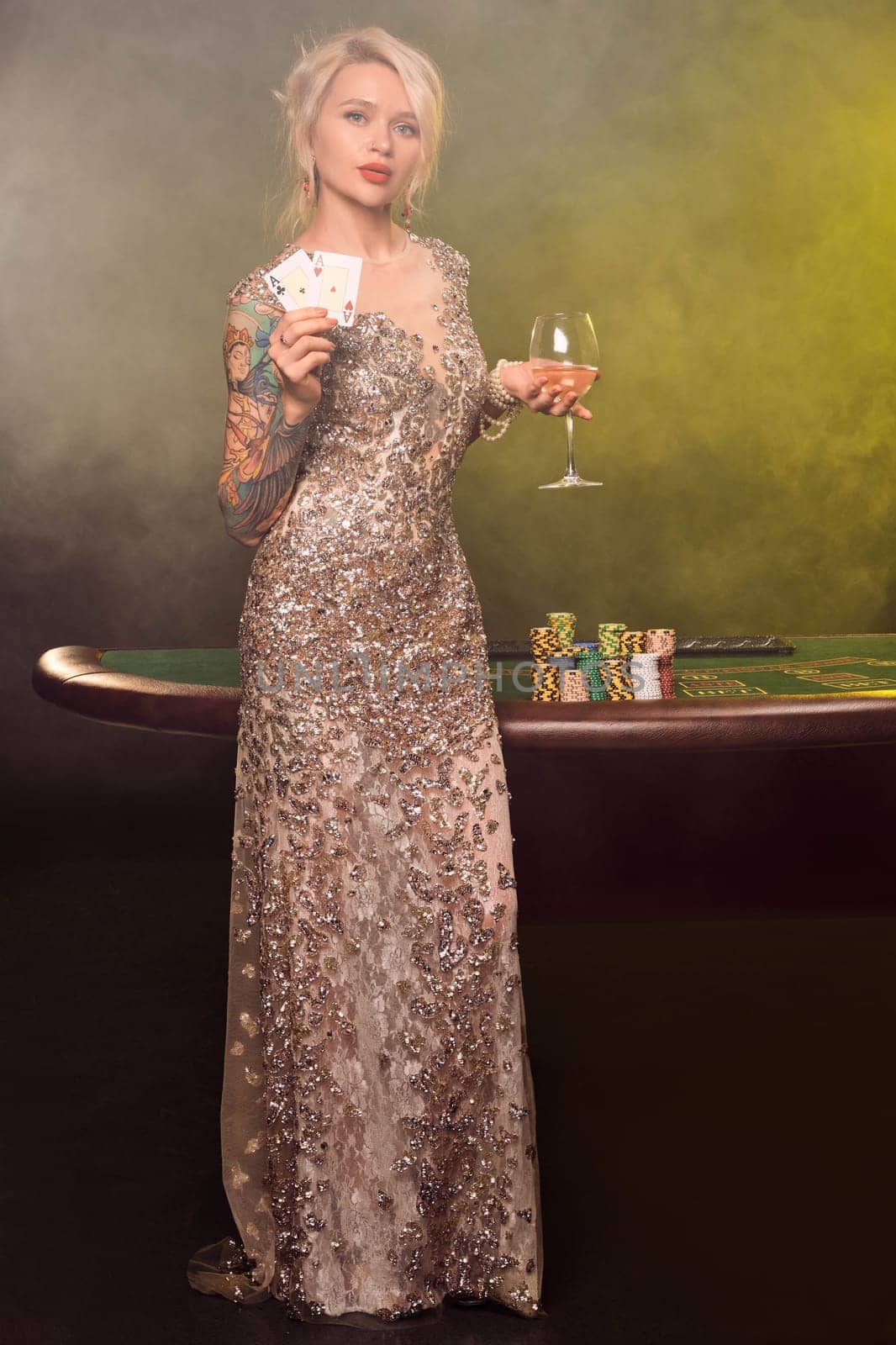 Full length shot of a lovely woman with blond hair, tattoed hands and perfect make-up, dressed in a silver shiny dress. She is standing against a gambling table, with two playing cards in her hands and smiling. Poker concept on a black smoke background with yellow and white backlights. Casino.