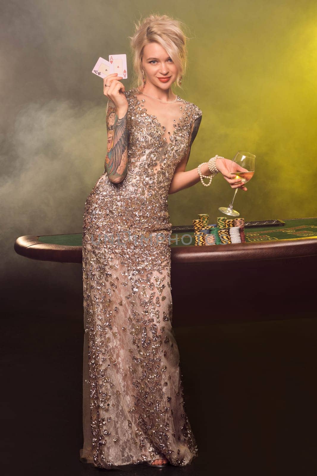 Full length shot of a cute lady with blond hair, tattoed hands and perfect make-up, dressed in a silver shiny dress. She is standing against a gambling table, with with a glass of champagne and two playing cards in her hands and smiling. Poker concept on a black smoke background with yellow and white backlights. Casino.