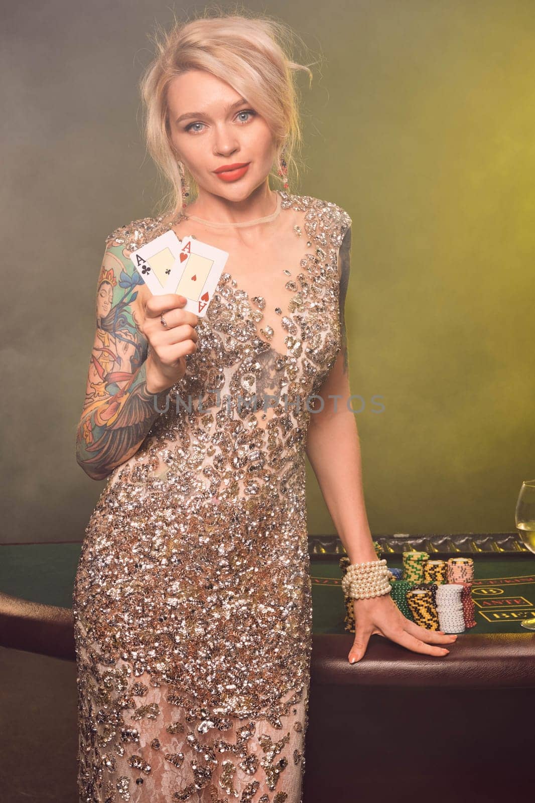 Close-up shot of a cute girl with blond hair, tattoed hands and perfect make-up, dressed in a silver shiny dress. She is standing against a gambling table, with two playing cards in her hands and looking at the camera. Poker concept on a black smoke background with yellow and white backlights. Casino.