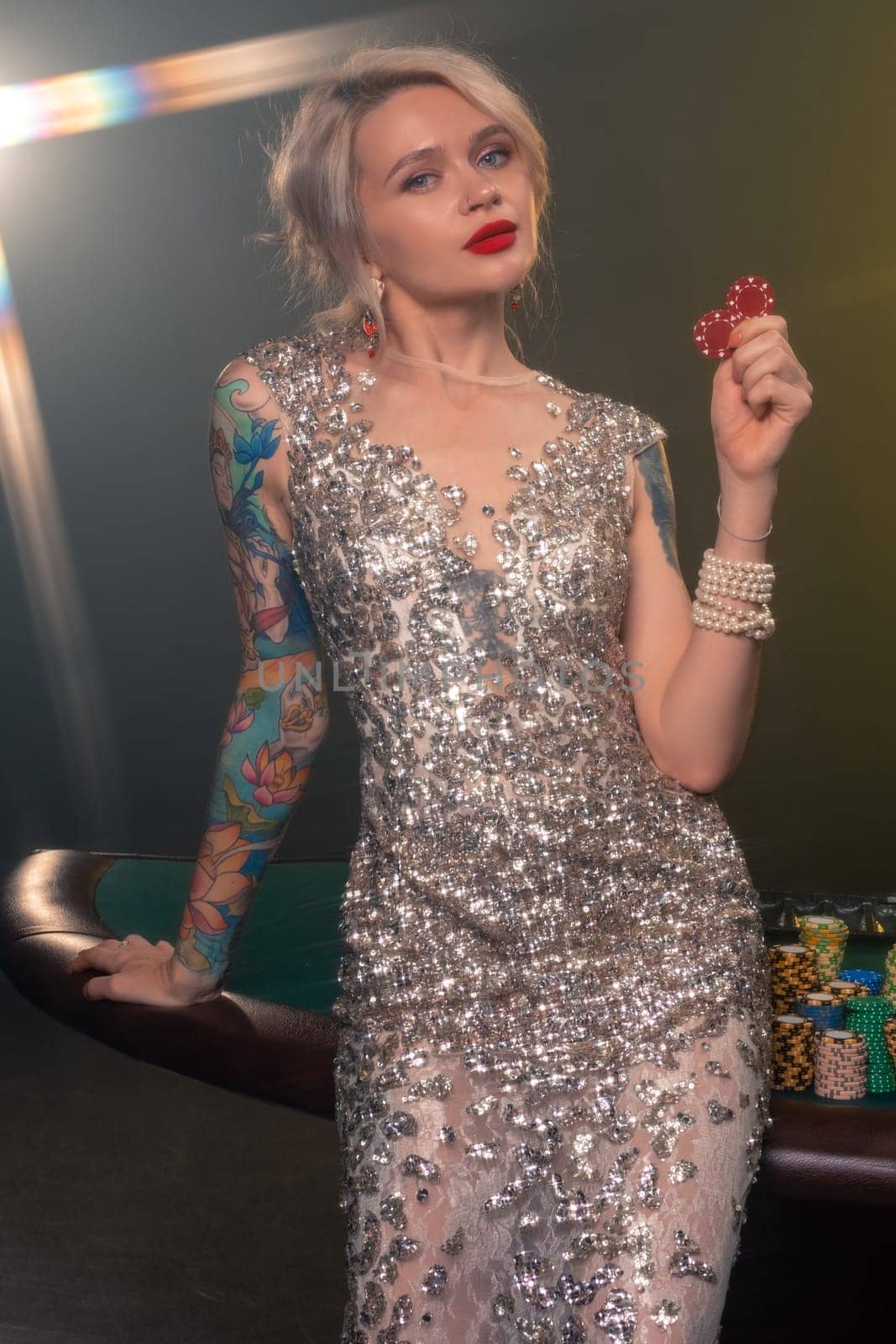 Close-up shot of an graceful maiden with blond hair, tattoed hands and perfect make-up, dressed in a silver shiny dress. She is standing against a gambling table, with red chips in her hands and looking at the camera. Poker concept on a black smoke background with a backlight. Casino.
