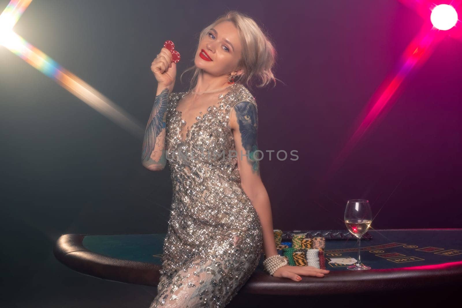 Medium close-up shot of a graceful female with blond hair, tattoed hands and perfect make-up, dressed in a silver shiny dress. She is standing against a gambling table, with red chips in her hands and smiling. Poker concept on a black smoke background with pink and blue backlights. Casino.