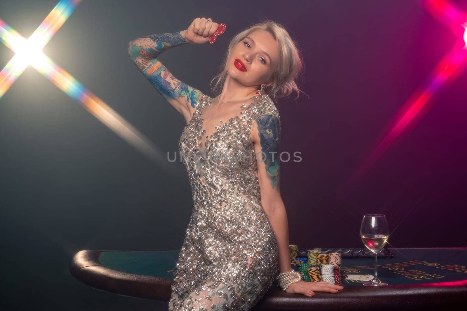 Medium close-up shot of a graceful woman with blond hair, tattoed hands and perfect make-up, dressed in a silver shiny dress. She is standing against a gambling table, with red chips in her hands and looking at you. Poker concept on a black smoke background with pink and blue backlights. Casino.