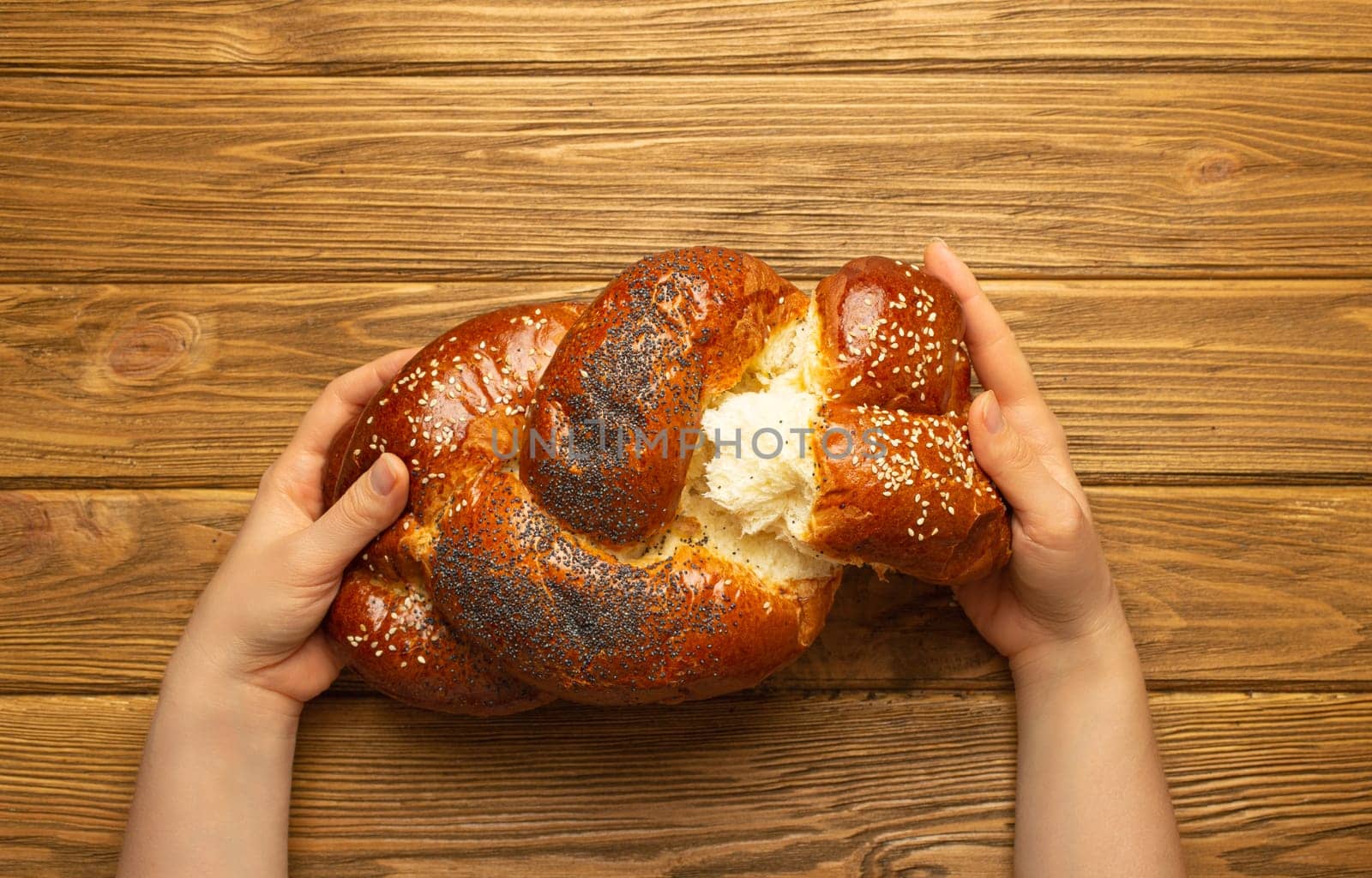 Female hands tearing off a piece of freshly baked Challah bread covered with poppy and sesame seeds, top view on rustic wooden background, traditional festive Jewish cuisine by its_al_dente