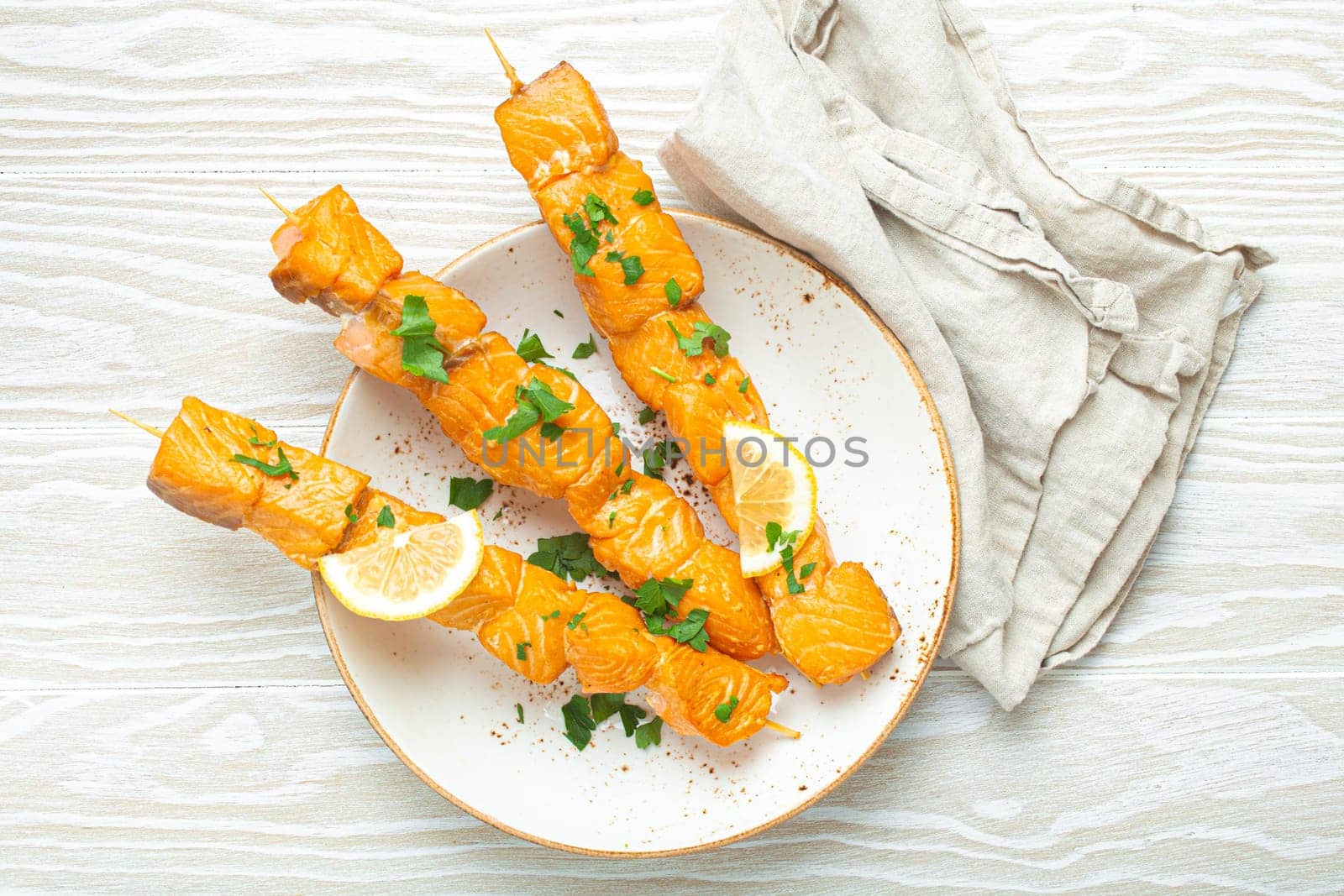 Grilled barbecue salmon skewers seasoned with green parsley and lemon on ceramic plate on white wooden rustic table background top view, healthy eating.