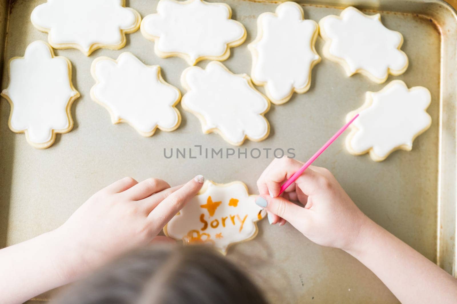 A heartwarming scene of a little girl carefully writing 'Sorry' on sugar cookies with food coloring, the cookies beautifully flooded with white royal icing.