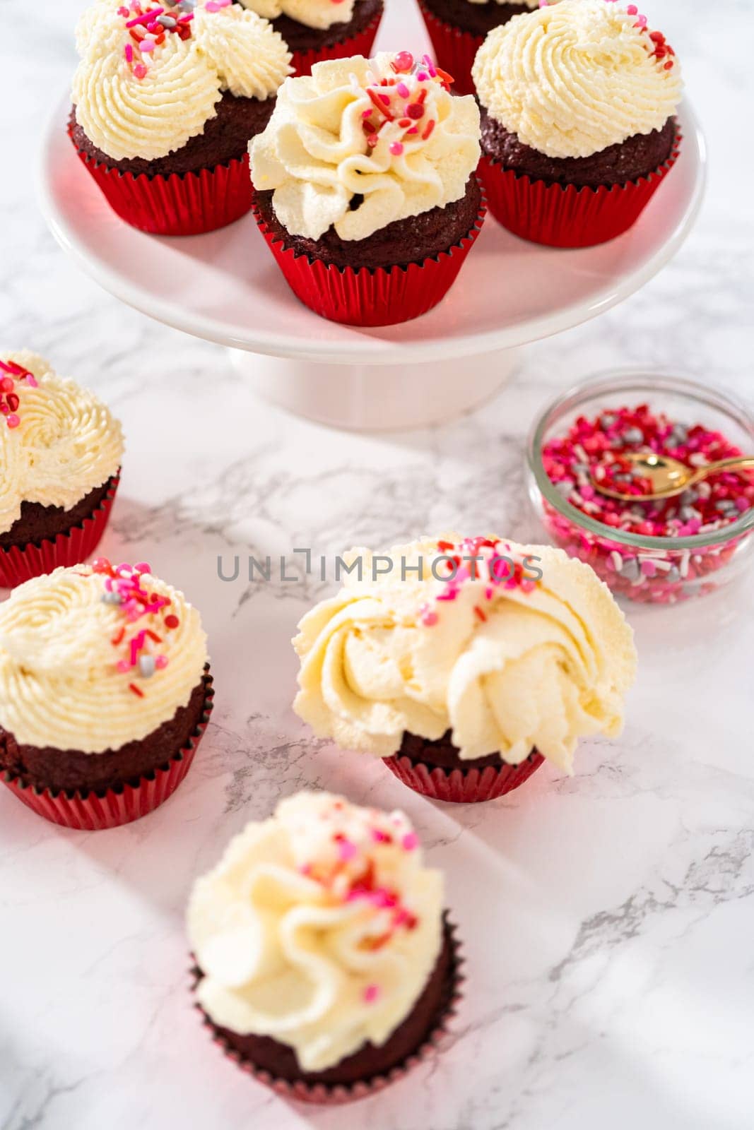 Freshly baked velvet cupcakes with white chocolate ganache frosting decorated with sprinkles.