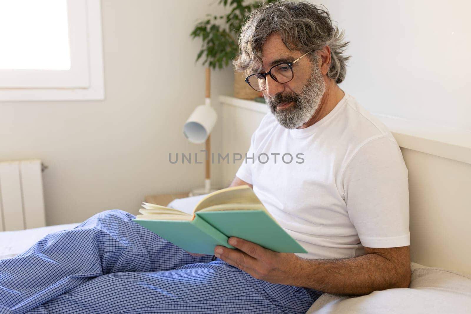Mature man with glasses relaxing in bed, reading a book wearing pyjamas. Lifestyle concept.