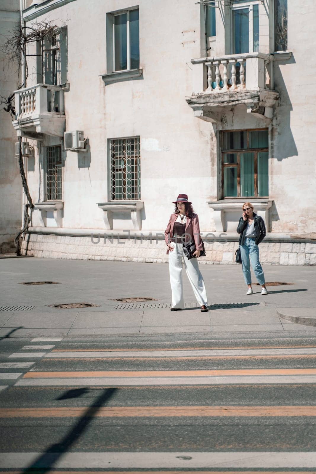 Woman city road crossing. Stylish woman in a hat crosses the road at a pedestrian crossing in the city. Dressed in white trousers and a jacket with a bag in her hands