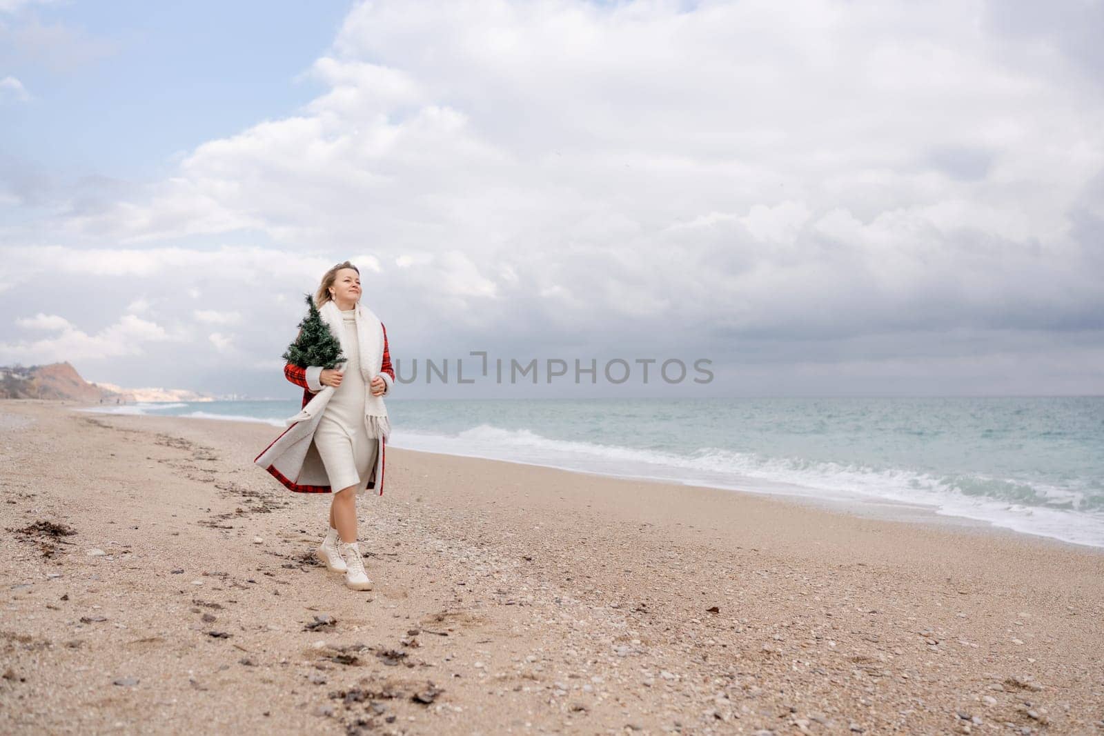 Blond woman holding Christmas tree by the sea. Christmas portrait of a happy woman walking along the beach and holding a Christmas tree in her hands. Dressed in a red coat, white dress