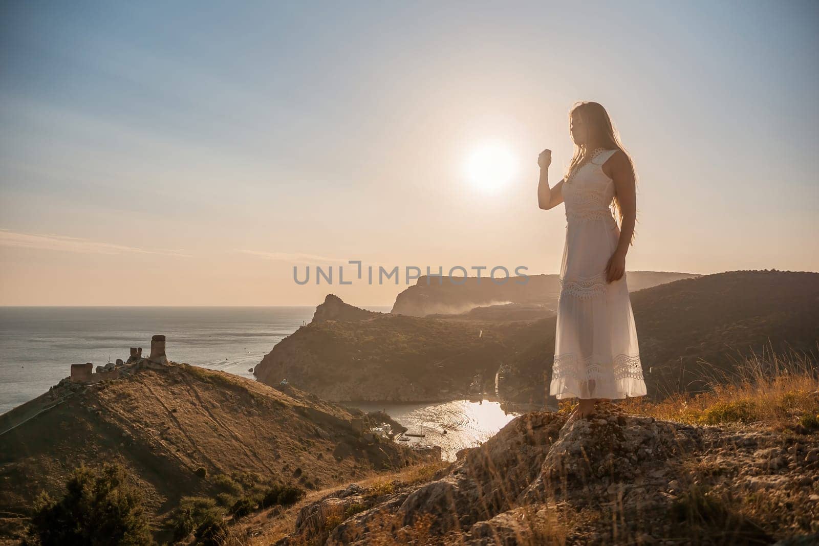 A woman stands on a rocky hill overlooking the ocean. She is wearing a white dress and she is enjoying the view. The scene is serene and peaceful, with the sun shining brightly in the background. by Matiunina