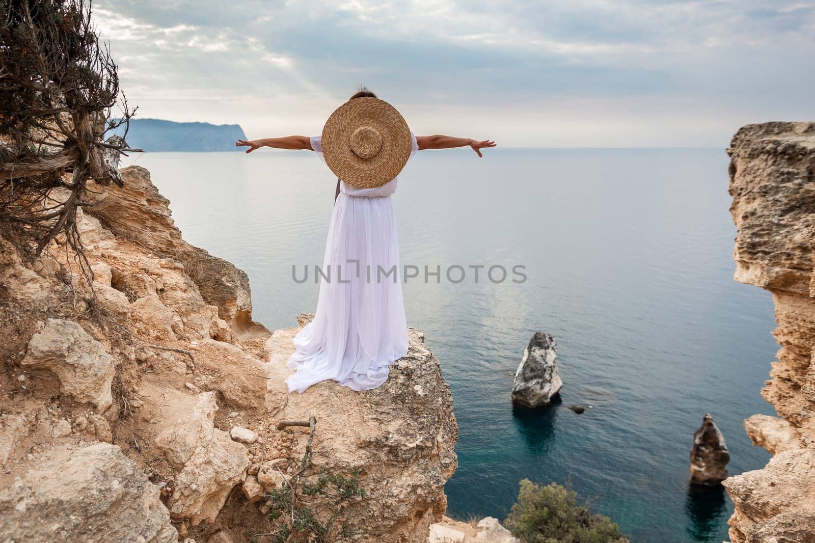woman white dress stands on a rocky cliff overlooking the ocean. She is wearing a straw hat and she is enjoying the view. by Matiunina