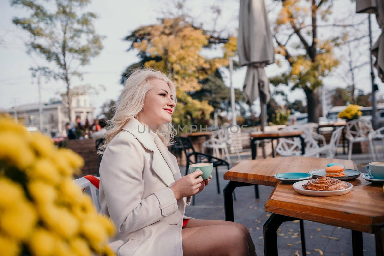 A woman is sitting at a table with a cup of coffee and a pastry. She is smiling and enjoying her time. by Matiunina