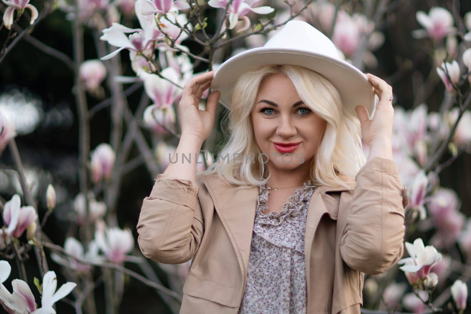 Magnolia flowers woman. A blonde woman wearing a white hat stands in front of a tree with pink flowers. She has a smile on her face and she is enjoying the beautiful scenery