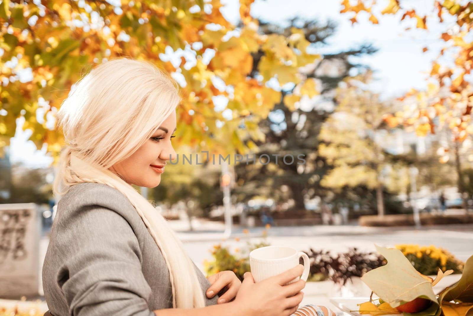 A blonde woman is sitting at a table with a cup of coffee. She is wearing a gray jacket and has a smile on her face. The scene takes place in a park with trees in the background