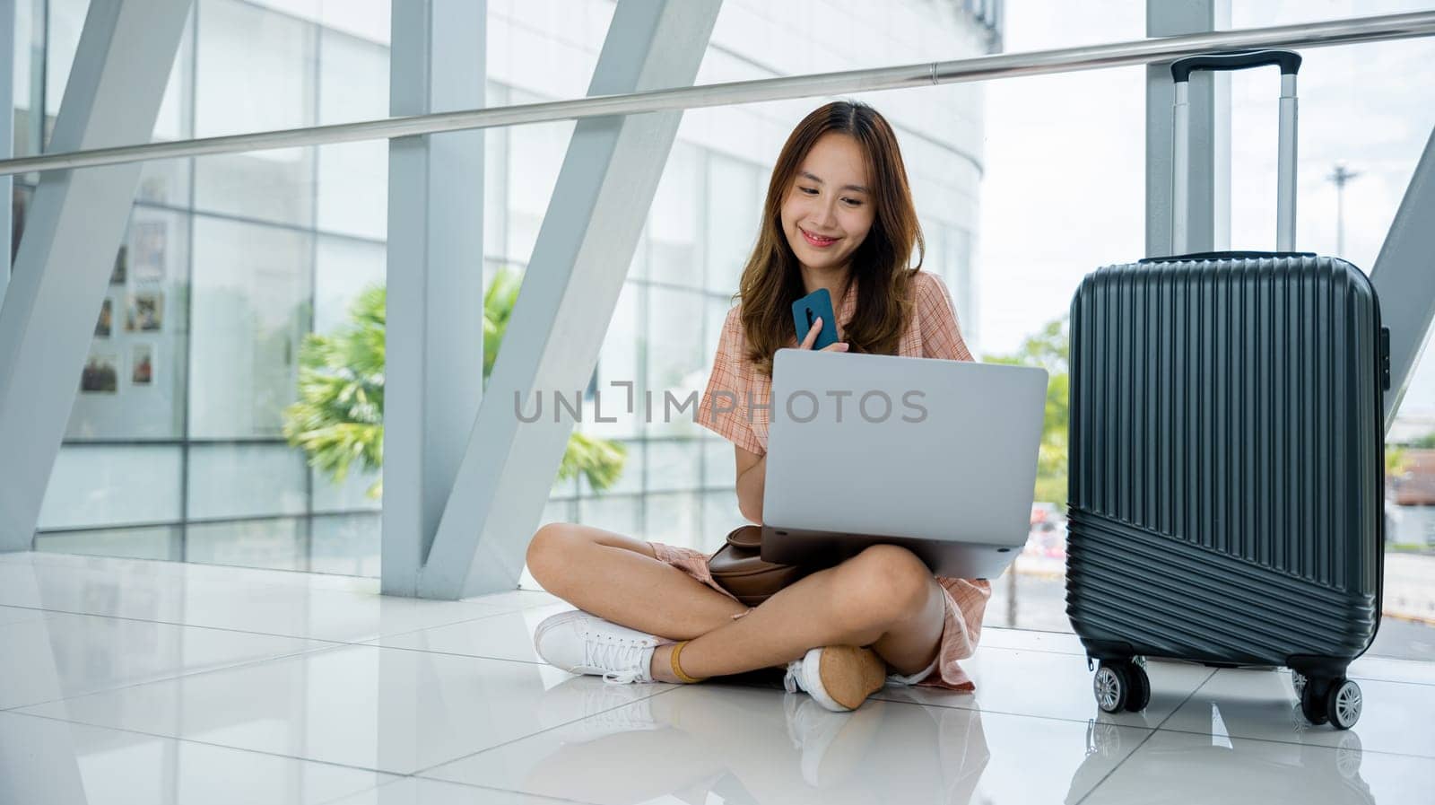 A girl sitting on the airport floor, working on her phone and laptop, with her suitcase nearby. She's a global traveler, always on the move. Multitasking is her workstyle. Air travel concept
