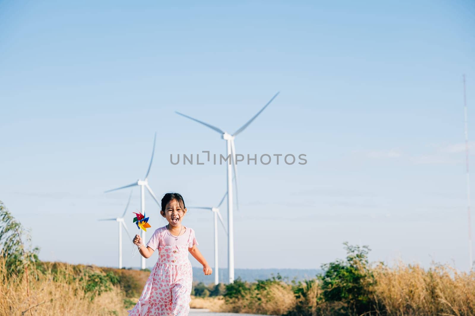 Little girl's playful interaction near windmills holding pinwheels while running. Embracing wind energy education promoting clean electricity and a cheerful childhood.