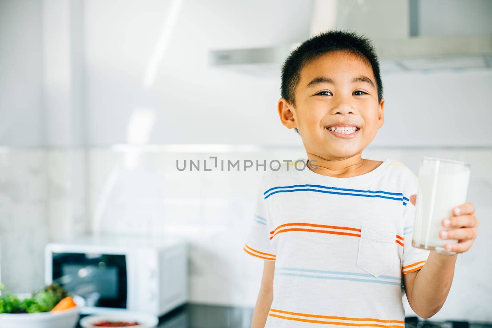 Portrait of cheerful Asian preschooler, boy holds milk in kitchen. Smiling son enjoys drink, radiating joy. Happy child sips calcium-rich liquid, feeling cheerful at home give me.