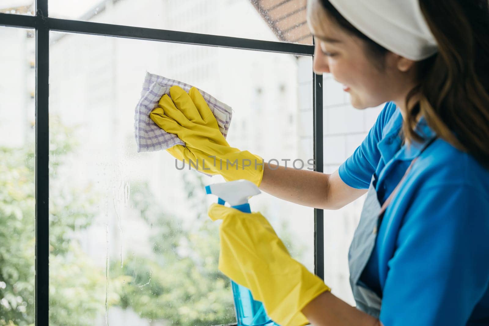 In an office setting a happy maid uses a spray to clean windows. Her housework routine ensures transparency using gloves and cleaning products for sparkling clean windows.