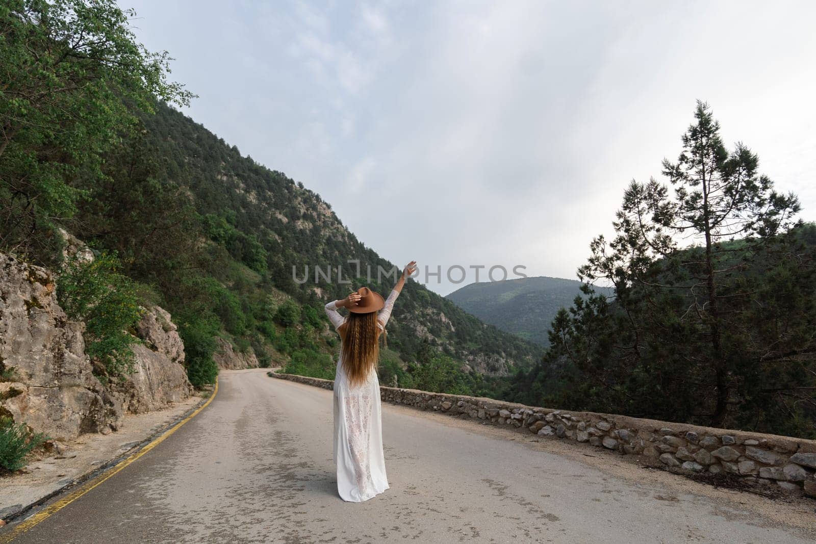 A woman in a white dress is standing on a road, with a hat on her head. The scene is set in a mountainous area, with trees in the background. The woman is enjoying the view and the fresh air