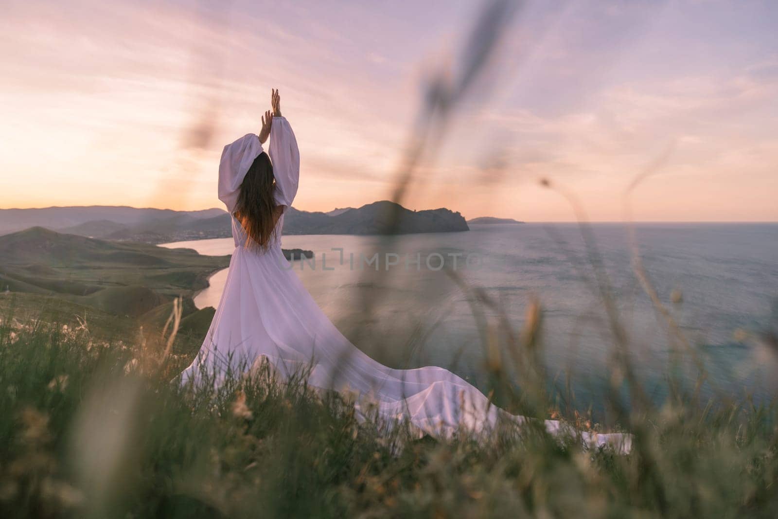 A woman in a white dress is standing on a grassy hill overlooking the ocean. She is in a yoga pose, with her arms raised above her head. The scene is serene and peaceful, with the ocean as a backdrop