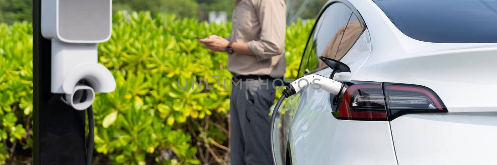 Young man use smartphone to pay for electricity at public EV car charging station green city park. Modern environmental and sustainable urban lifestyle with EV vehicle. Panorama Expedient