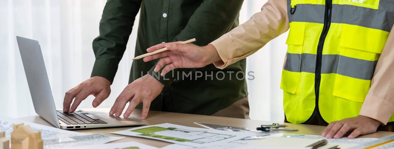 Architect and safety engineer discussion about building blueprint displaying on laptop on table with architectural document and wooden block scatter around at modern office. Closeup. Delineation.