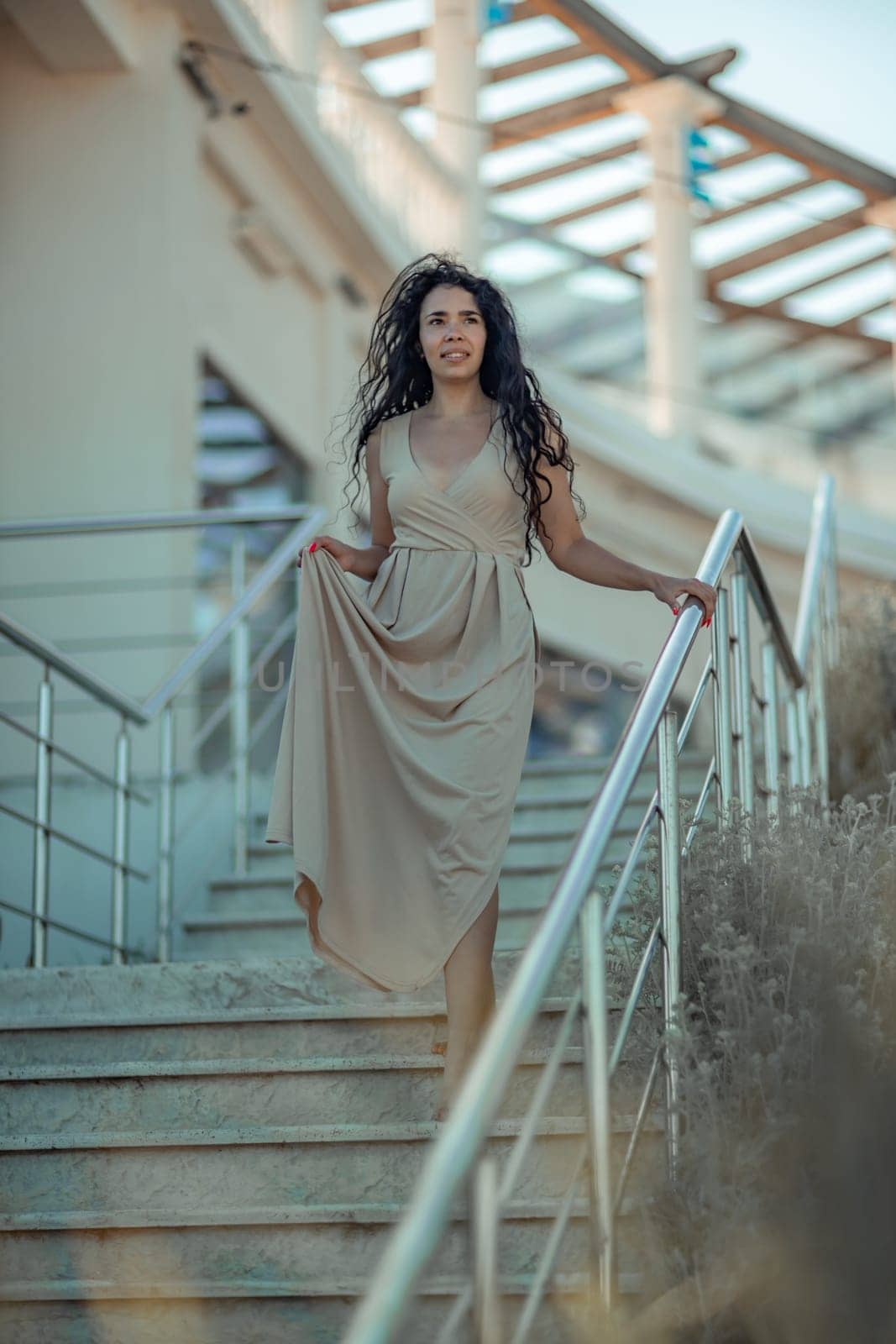 A woman in a dress stands on a set of stairs. The stairs are made of metal and are located in front of a building. The woman is wearing a necklace and a bracelet