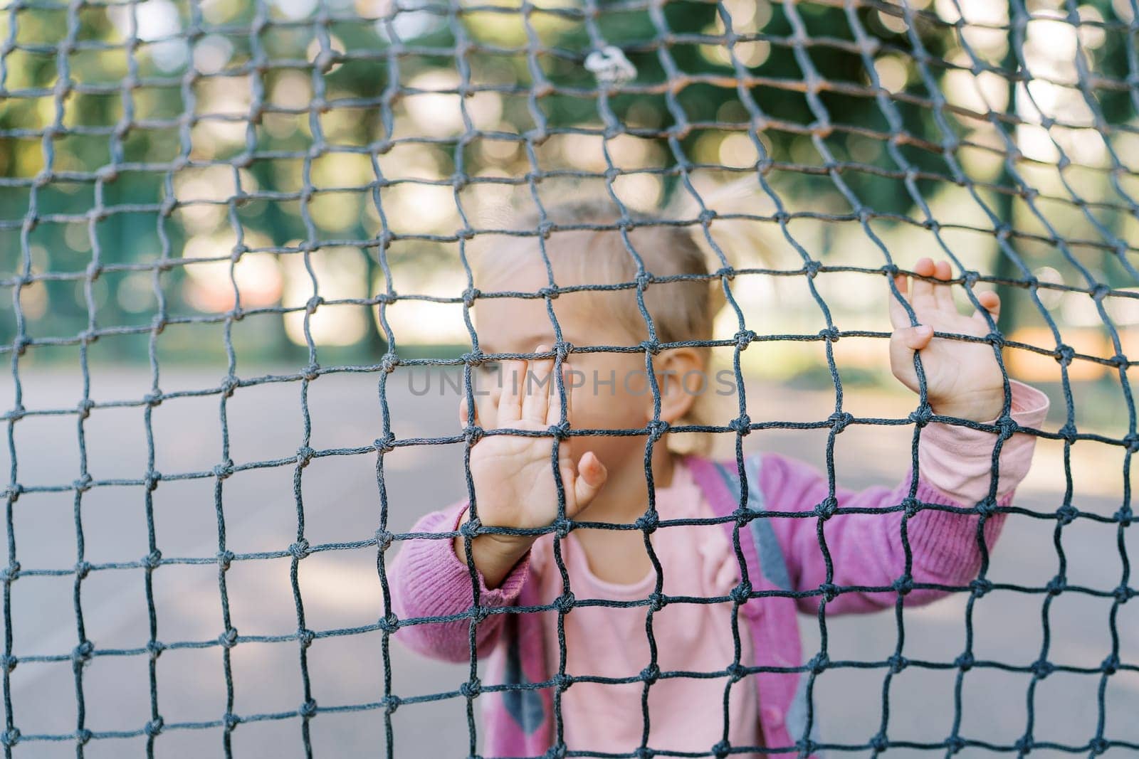 Little girl stands behind a tennis net and rests her hands on it. High quality photo