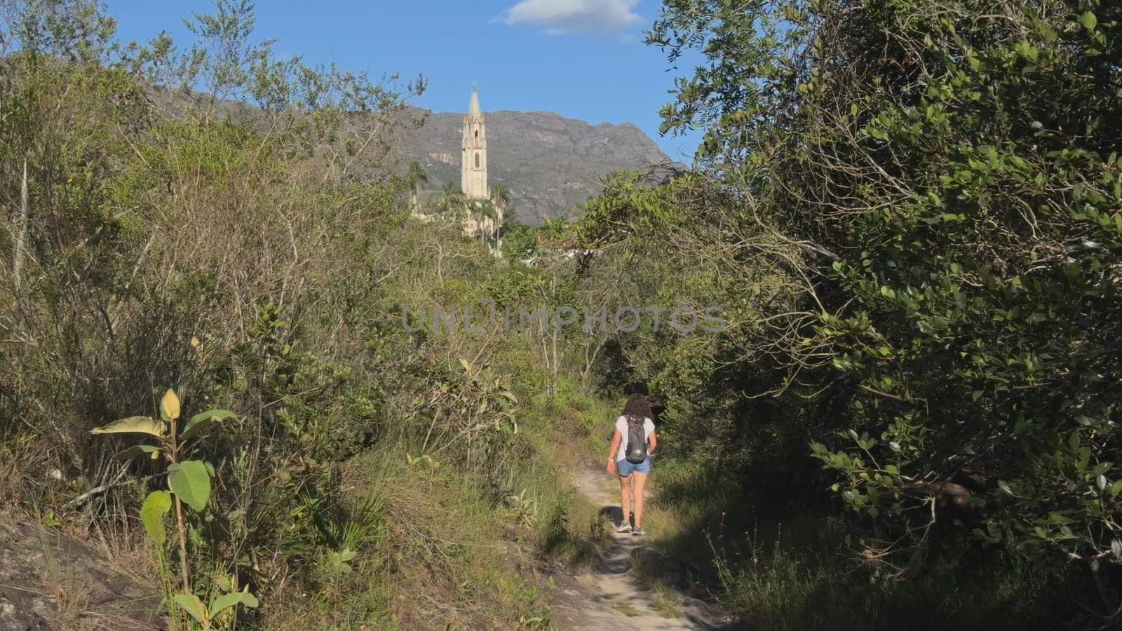 Hiker treks on forest path, church spire visible far.
