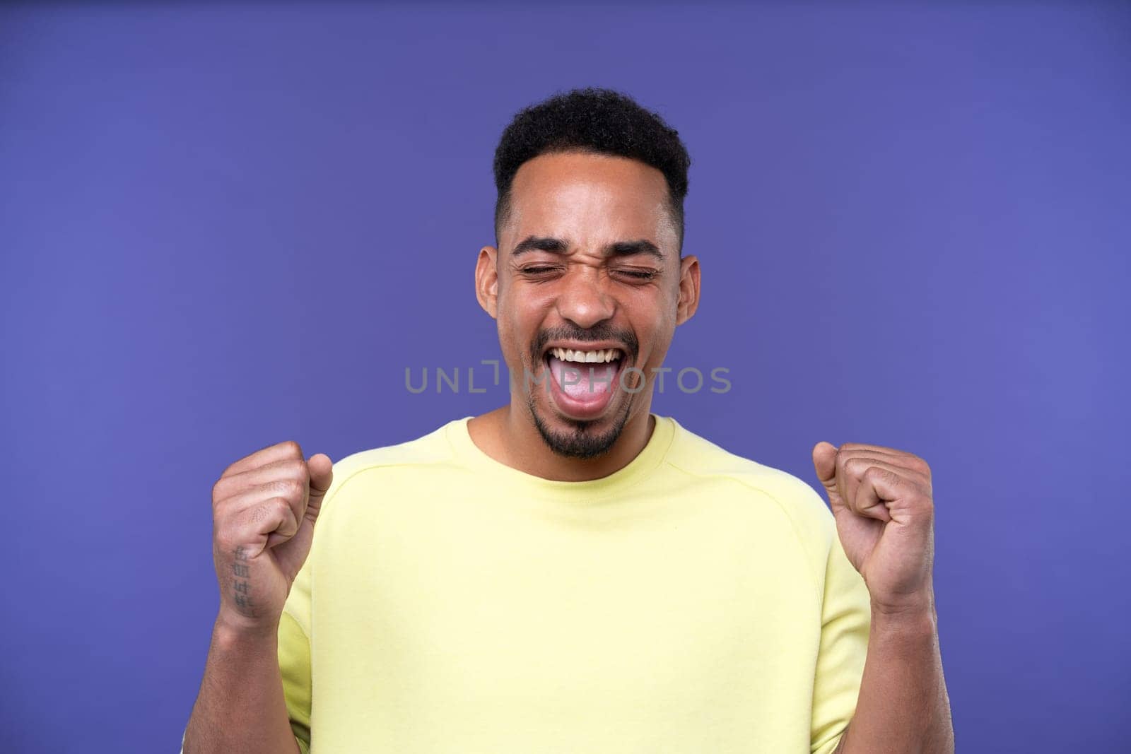 Emotional young black guy in casual raising fists up studio background, celebrating success, happy handsome african american millennial man sharing good emotions, copy space