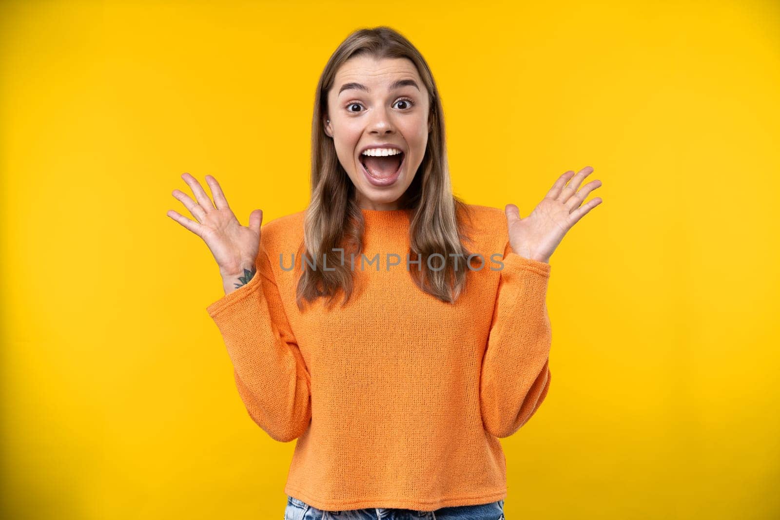 Happy emotions concept. Positive and beautiful young woman laughs poisitively looks aside with carefree face expression wears casual orange sweater isolated over yellow studio background.