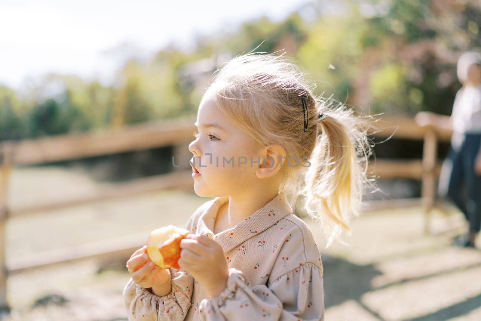 Little girl looks into the distance holding a bitten apple in her hands while standing in the park. High quality photo