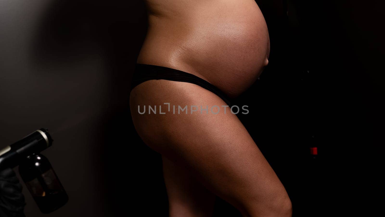 Faceless pregnant woman undergoing an instant tanning procedure. The master applies bronzer to the legs with a spray