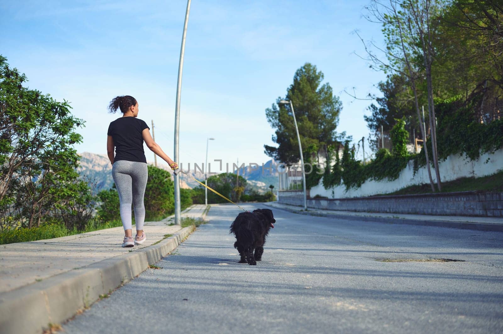 Rear view happy multi ethnic woman walking her dog on leash in the nature. Smiling female in sportswear, enjoying walk with her pedigree cocker spaniel outdoors. People and domestic animals concept