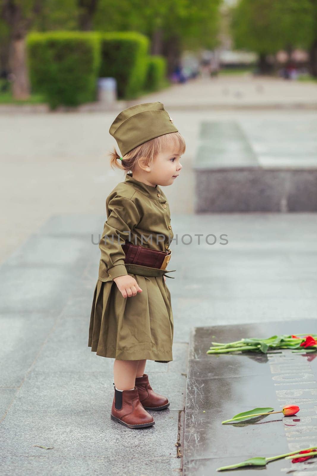 Adorable baby girl in Soviet military uniform lays a flower on a monument to those killed in World War II