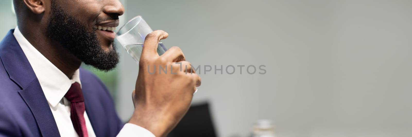 Unrecognizable bearded black businessman in blue formal suit holding glass of water, drinking water while working at office, panorama with copy space, cropped. Hydration, healthy lifestyle concept