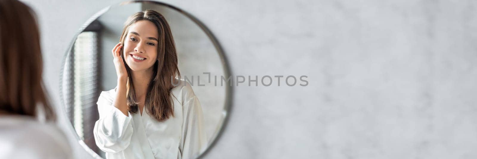 Beauty Concept. Portrait Of Attractive Happy Woman Looking At Mirror In Bathroom, Beautiful Millennial Lady Wearing White Silk Robe Smiling To Reflection, Enjoying Her Appearance, Copy Space