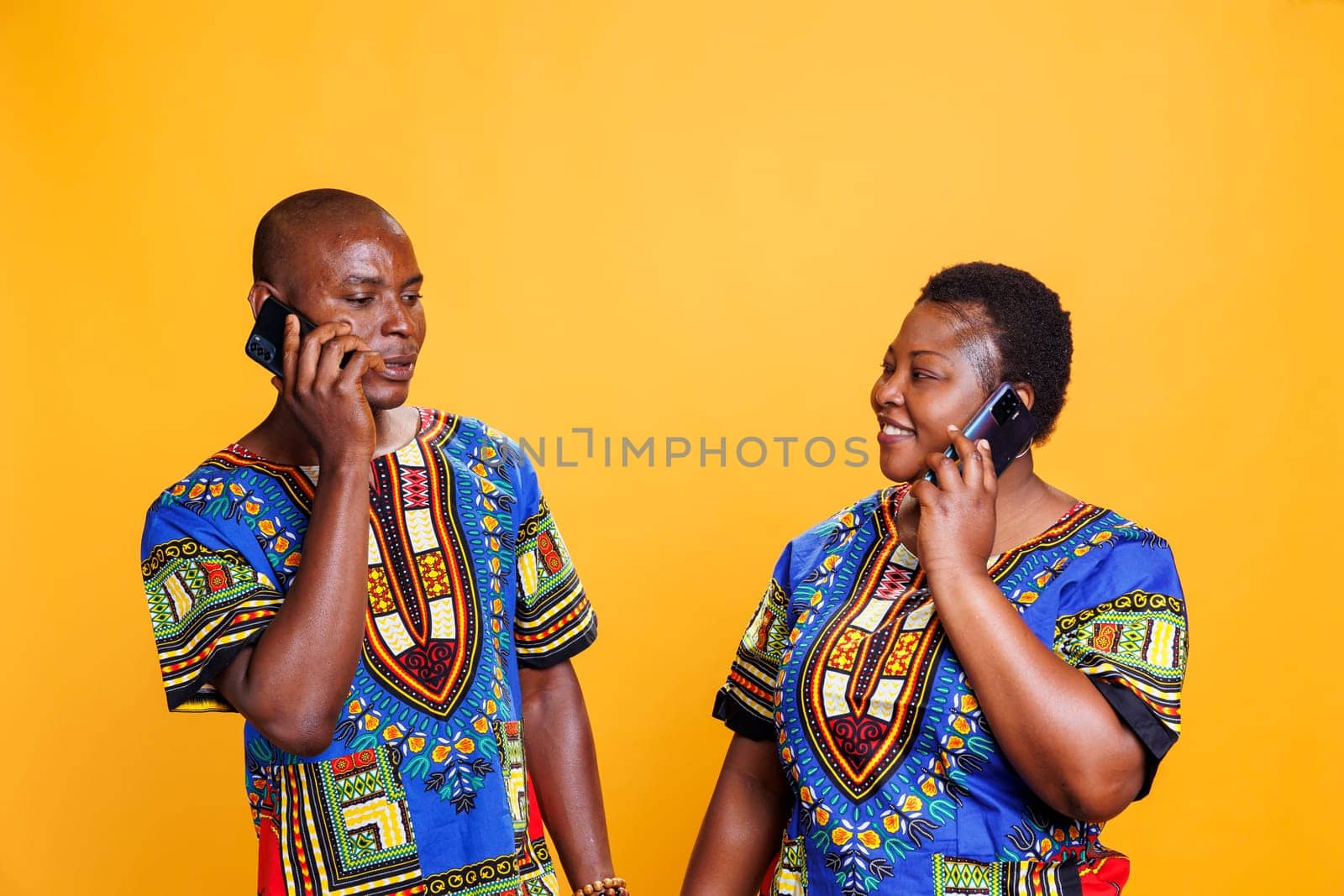 Smiling man and woman couple looking at each other while speaking on smartphone. Cheerful wife and husband having fun conversation on mobile phone while standing in studio