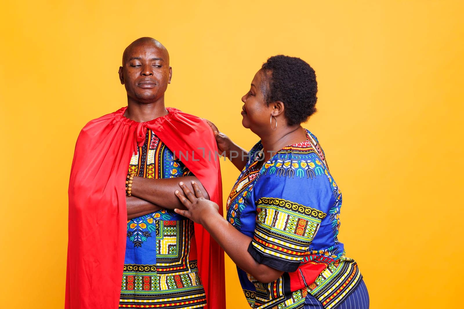 Happy wife appreciating husband in superhero cape, showing love and support. Confident african american man wearing red superman cloak standing near woman on studio background