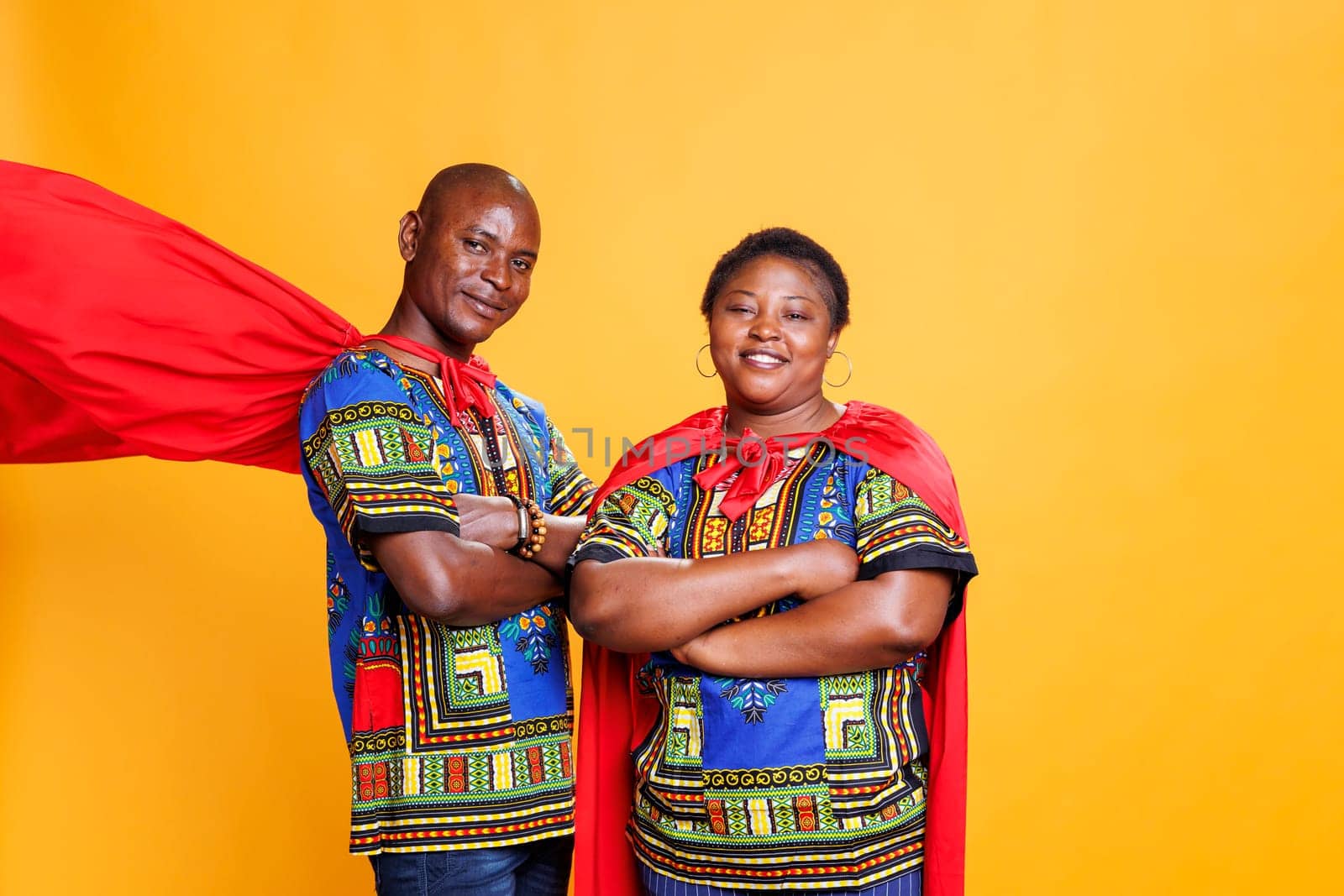 Smiling black couple dressed in superhero red cloaks posing with folded hands and looking at camera. Cheerful superman and superwoman pair wearing capes showing power and strength portrait