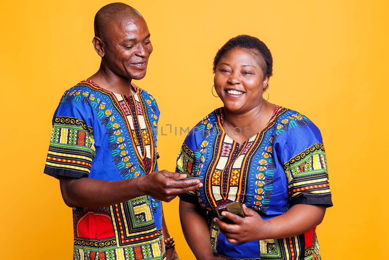 Smiling african american husband offering credit card to wife to pay online in internet store on smartphone. Woman laughing and looking at camera while shopping online with boyfriend