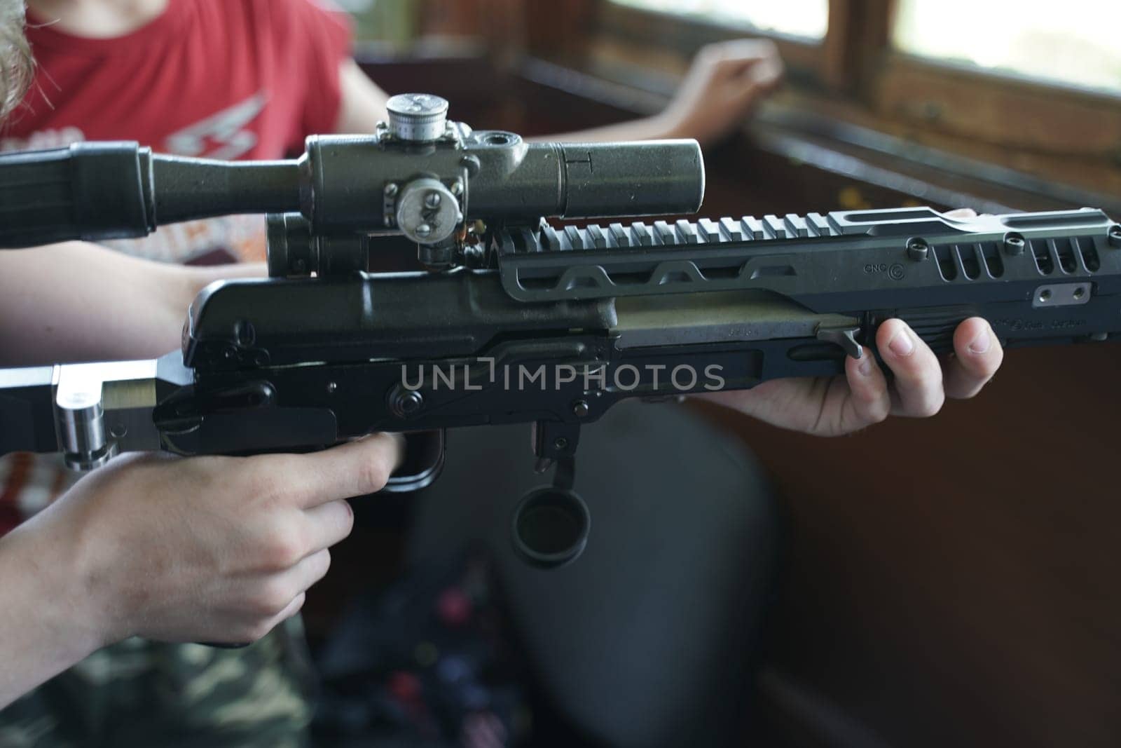 A child in camouflage clothing is holding a rifle equipped with an optical sight, focusing intently on handling the firearm.