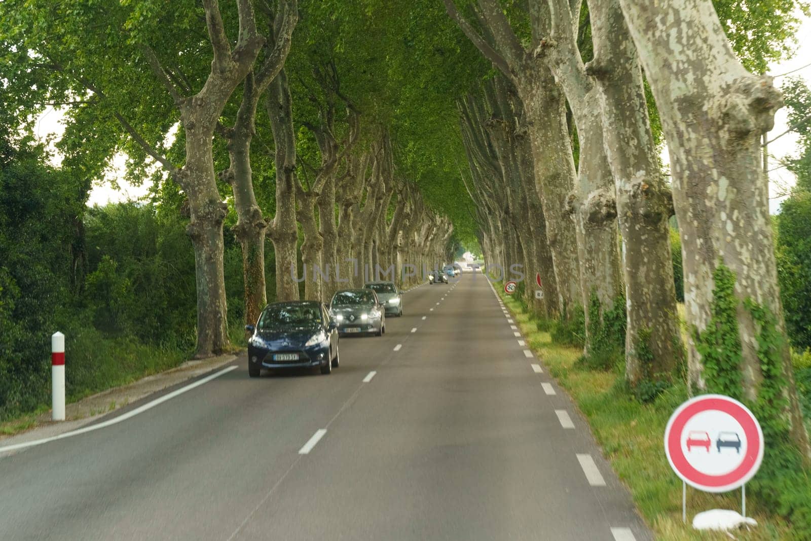 Nimes, France - May 30, 2023: Cars drive along a peaceful road flanked by tall, leafy trees, under a soft daylight, adhering to the no overtaking road sign.