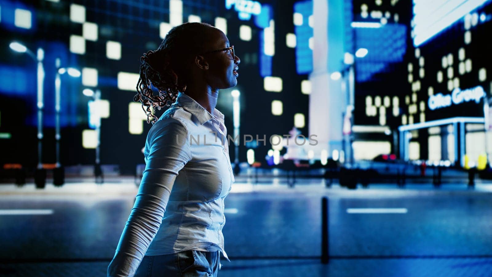 Smiling african american woman wandering around city boulevards during nighttime, pointing at interesting billboards on skyscrapers. Businesswoman strolling around streets illuminated by lamps