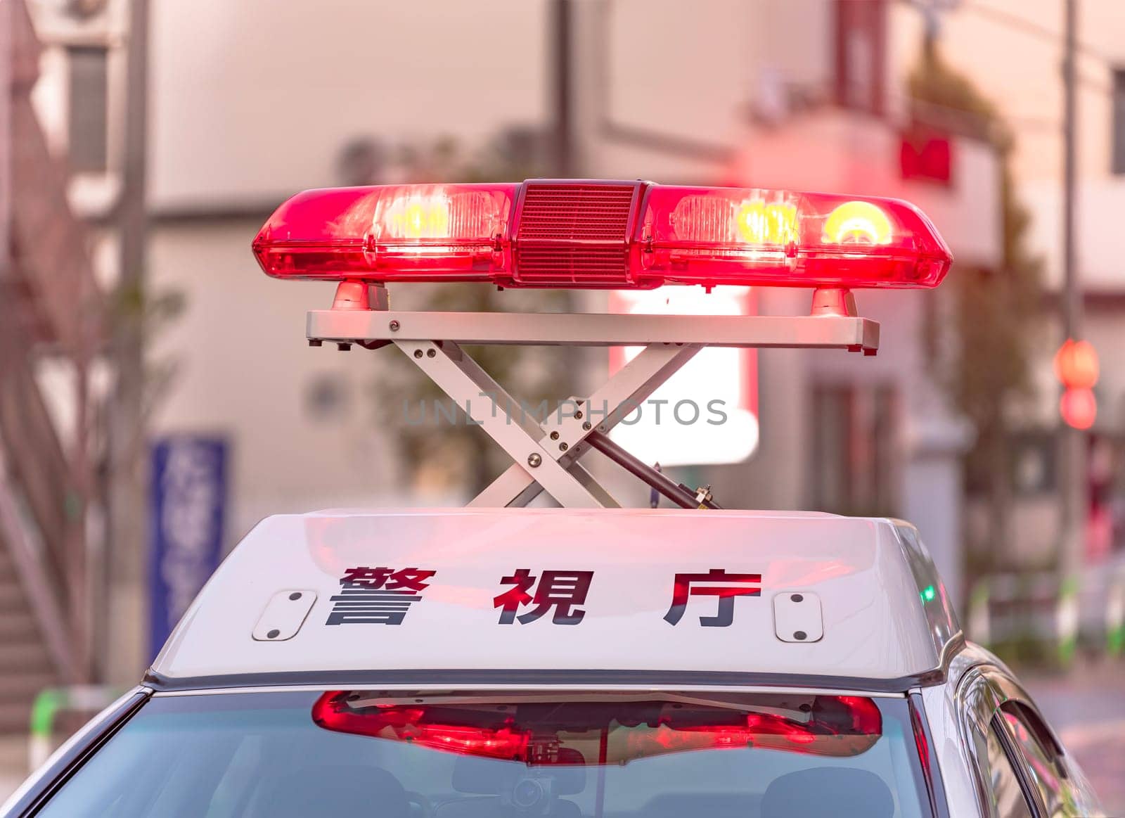 Close-up on bright red emergency beacons casting an intense glow as they swirl unfolded above the white roof of a Japanese police vehicle showing the word 'Keishicho' meaning 'Police'.