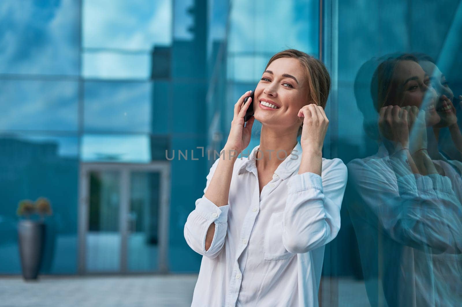 Businesswoman using smartphone outdoor. Business person talking mobile phone dressed white shirt standing outside near office building with glasses wall