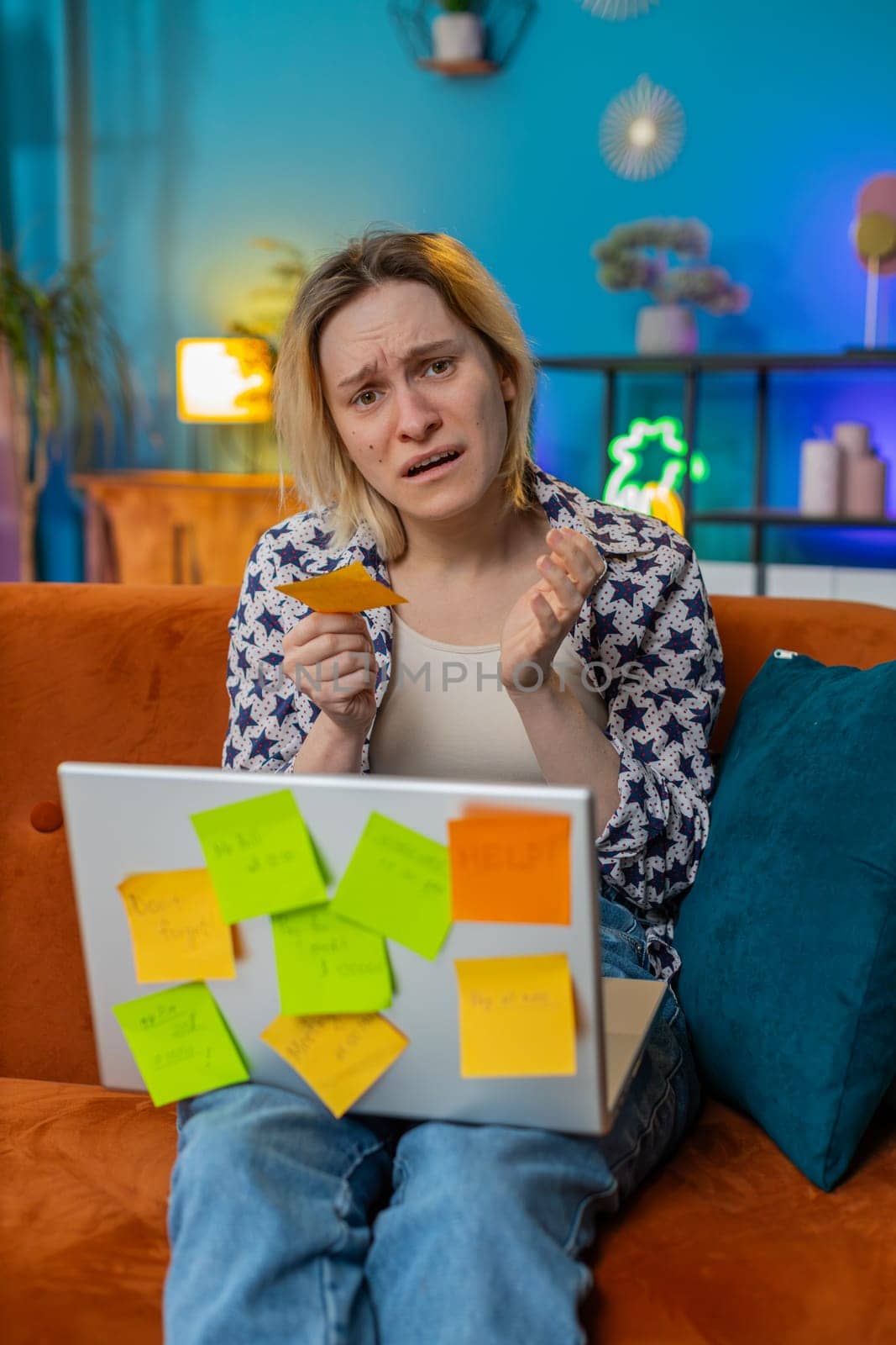 Tired exhausted young woman freelancer working on laptop sitting on sofa with many sticker task inscriptions. Frustrated girl busy feels sad boredom overworked multitasking. Panic attack, deadline.