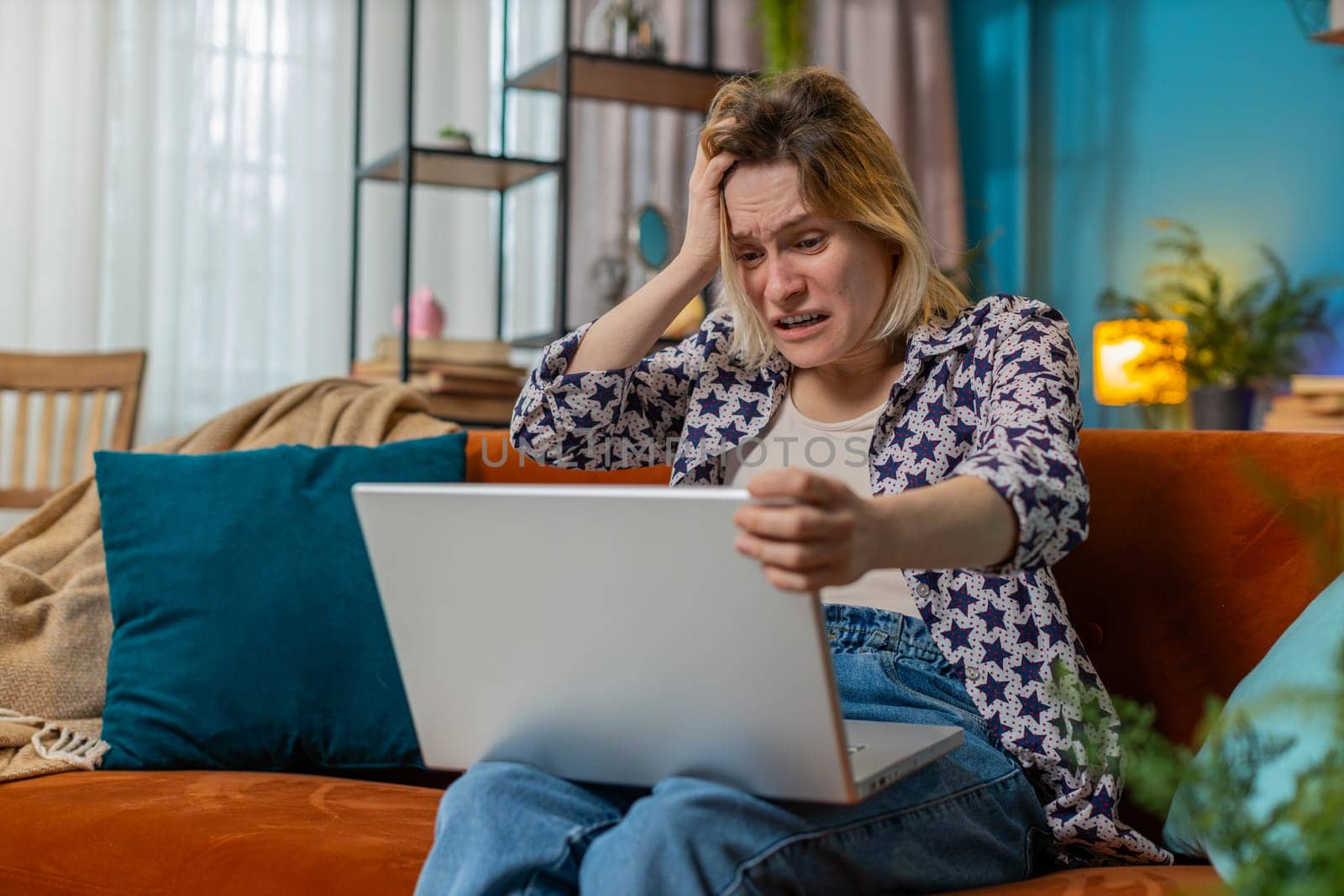 Displeased Caucasian woman using laptop notebook typing browsing working, loses becoming surprised sudden lottery results bad news fortune loss game fail computer virus. Young girl at home on couch.