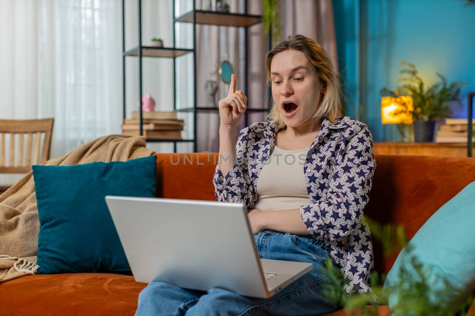 Thoughtful young Caucasian woman freelancer got an idea and typing on laptop netbook. Happy girl in casual clothes clenching fists and celebrating victory sitting on sofa in living room at home.