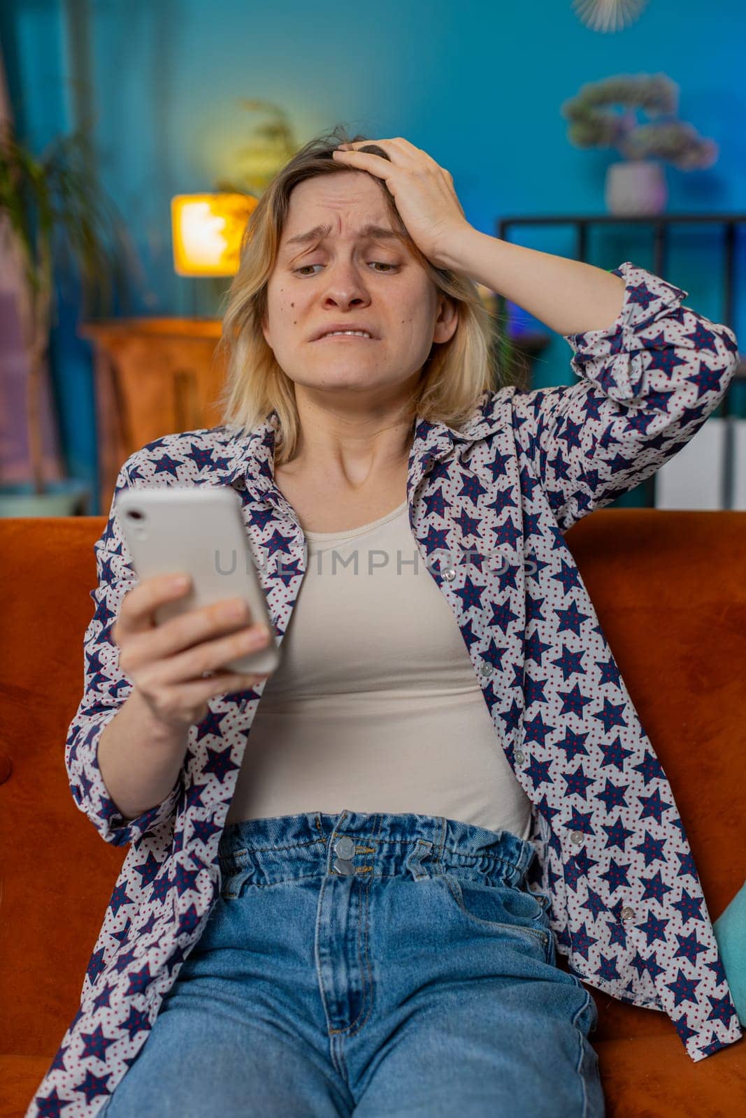 Shocked sad amazed woman using smartphone reading negative message feels annoyed sitting on couch in living room at home. Upset Caucasian girl having gadget trouble in apartment. App crash concept